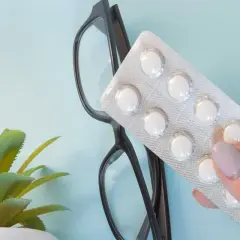 Una mano tiene un blister di pillole bianche, forse un'aspirina per le piante, accanto a un paio di occhiali e una piccola pianta verde.