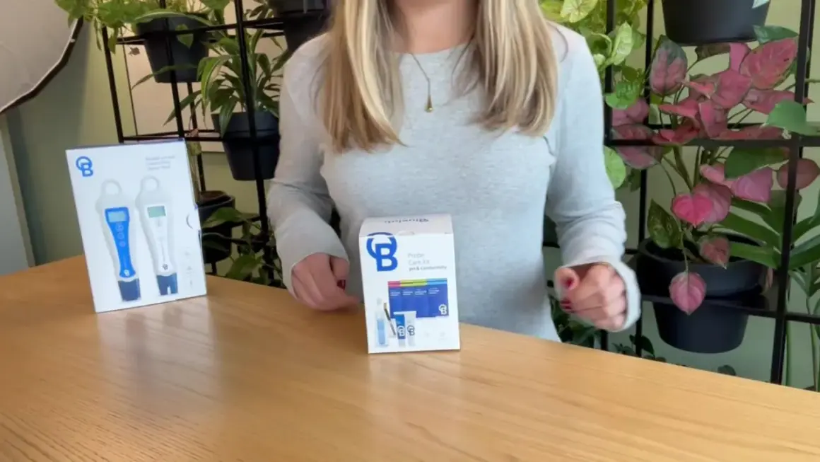 Kit di cura per sonde BlueLab con misuratori pH e conducibilità, su un tavolo di legno con piante sullo sfondo.