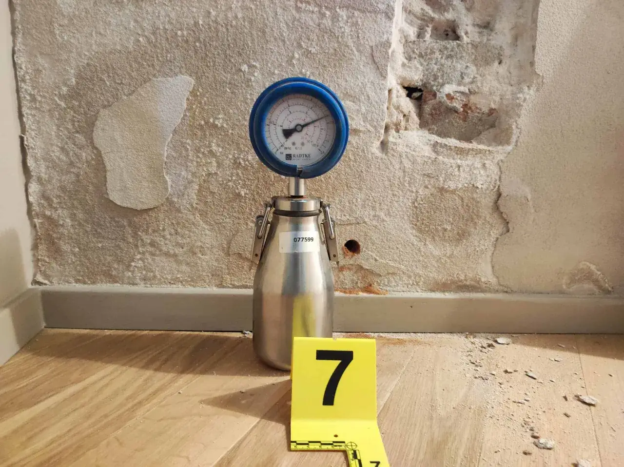 Misurazione umidità 70 per cento in casa. Strumento di misurazione su un contenitore metallico, accanto a un cartello giallo con il numero 7. Parete scrostata e pavimento in legno.